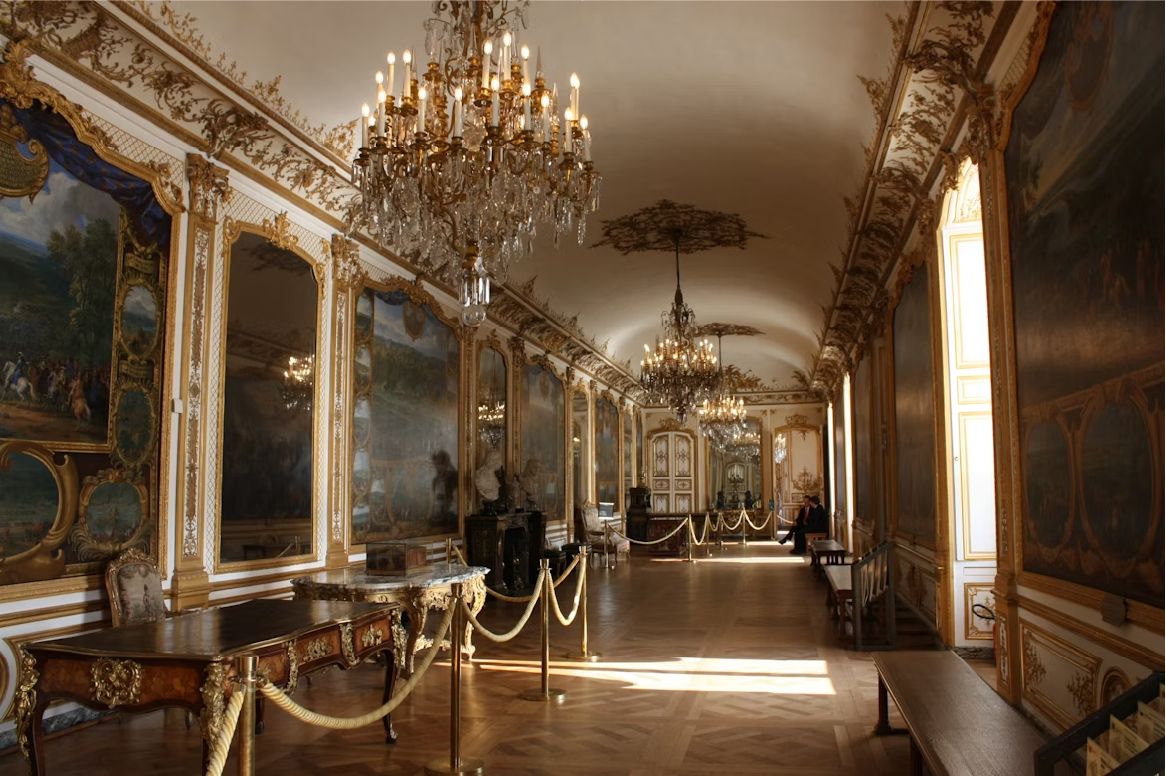 A long gilded hall with chandeliers and oil paintings