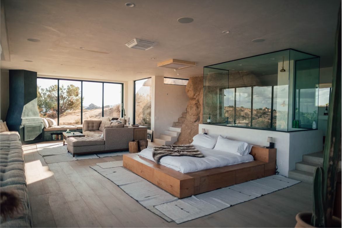 A wide bedroom with a low wooden bed, a glass bath and tall windows onto dry hills