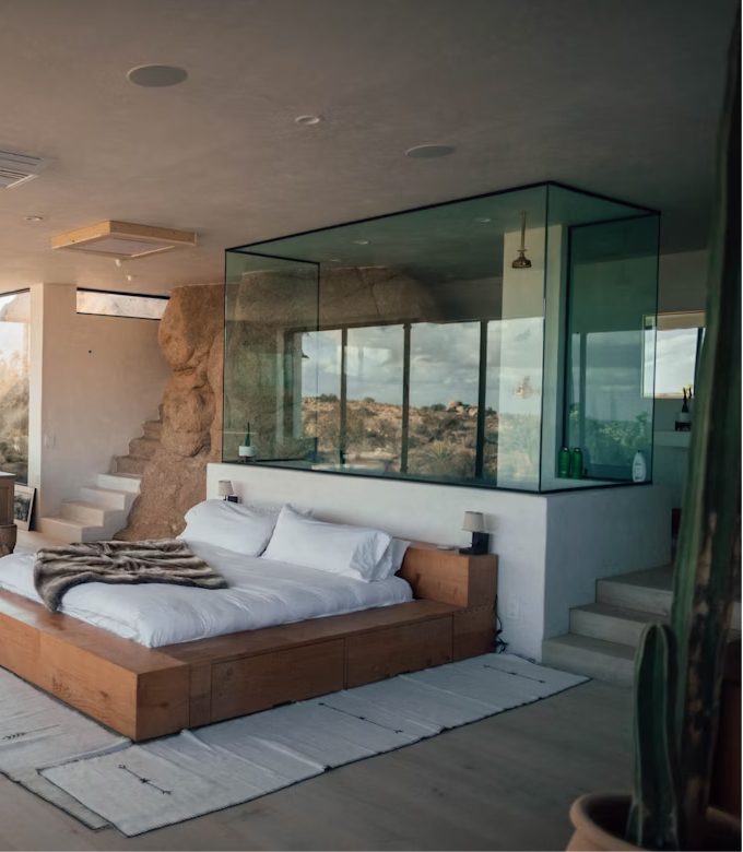 A glass bath box beside a bed, with a garden seen through the window